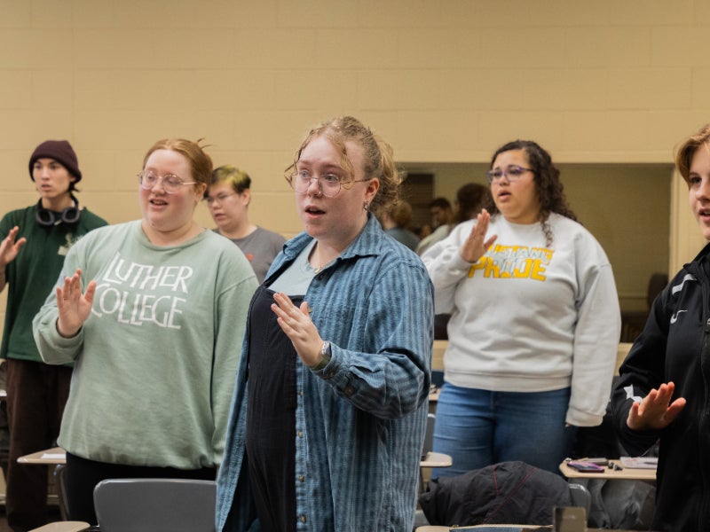 Students stand in a classroom, gesturing and singing