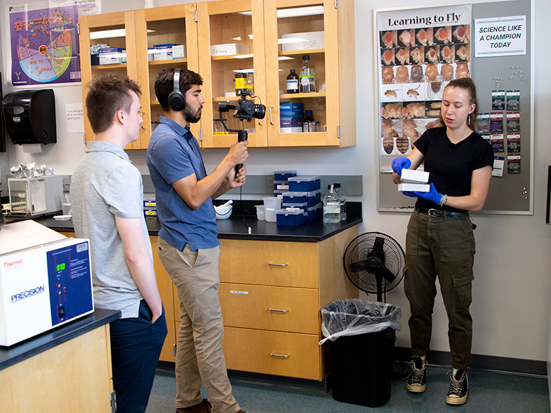 two Luther students filming a third Luther student presenting a science demonstration