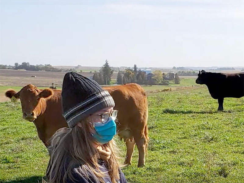 woman with long blonde hair, wearing a stocking cap, stands in a field with some cows