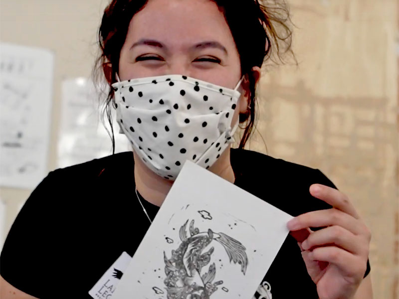 woman with black hair, wearing a mask, holds up a pen-and-ink drawing