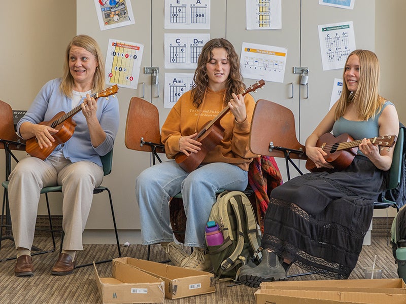 One professor and two students sit in a classroom, strumming ukuleles.