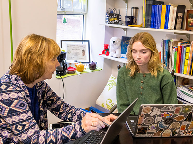 A professor and student sit together in conversation in a book-lined office with a window.