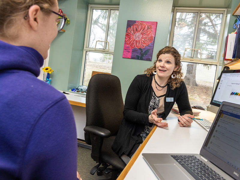 A professor and student sit in an office with windows to discuss academic progress