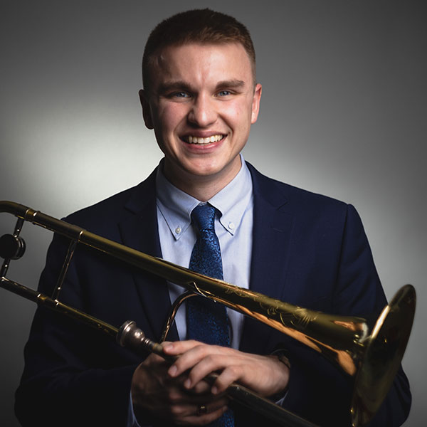 White man with short hair, smiling, wearing a jacket and tie, holding a trombone