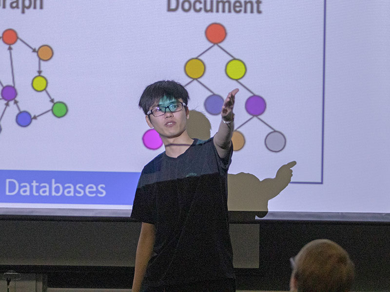 a man stands in front of a screen displaying the words "databases" and "document"; he is gesturing.