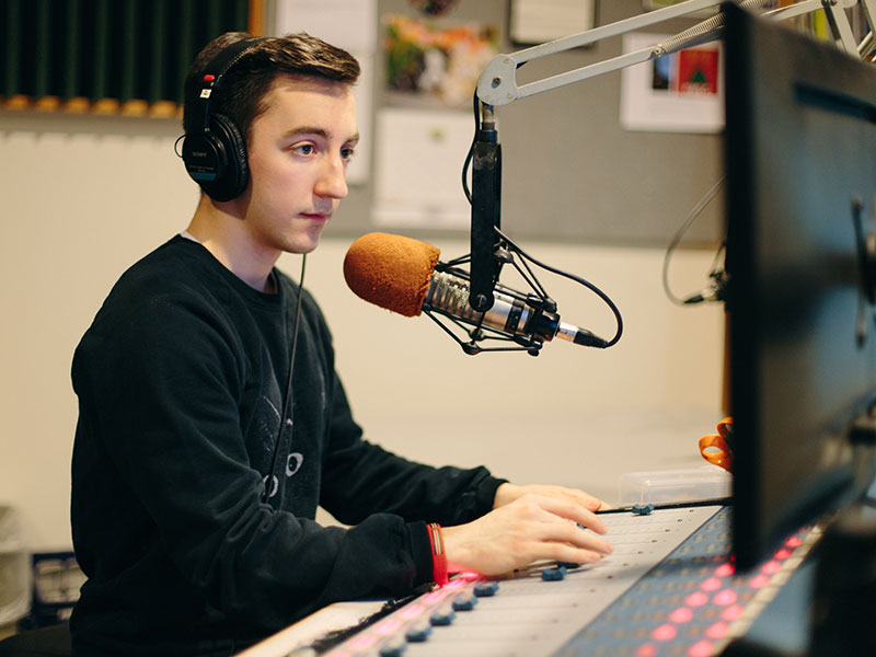 White man wearing headphones, sitting at a microphone and control board