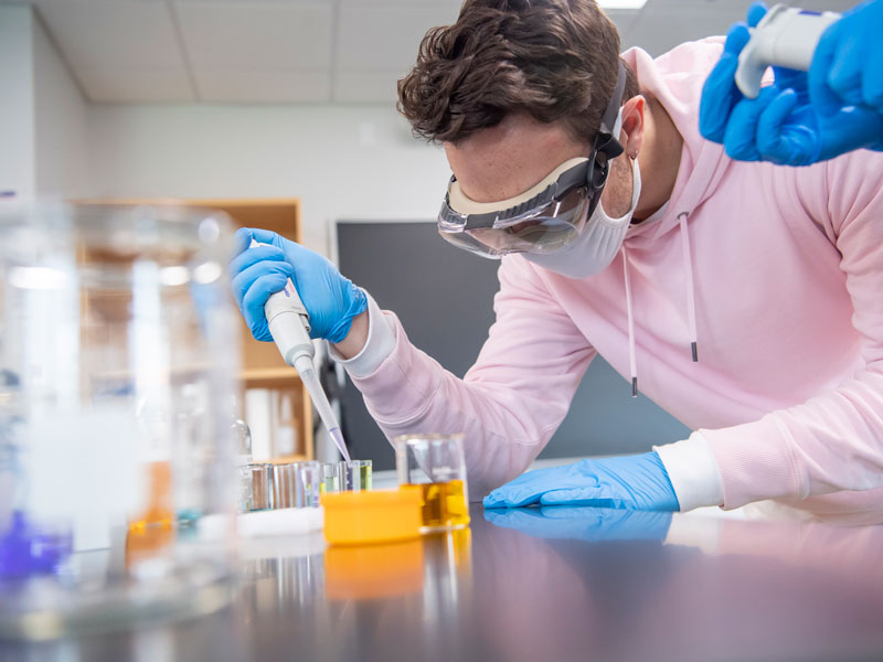 a male student wearing a gloves, goggles and mask, puts a solution into a beaker
