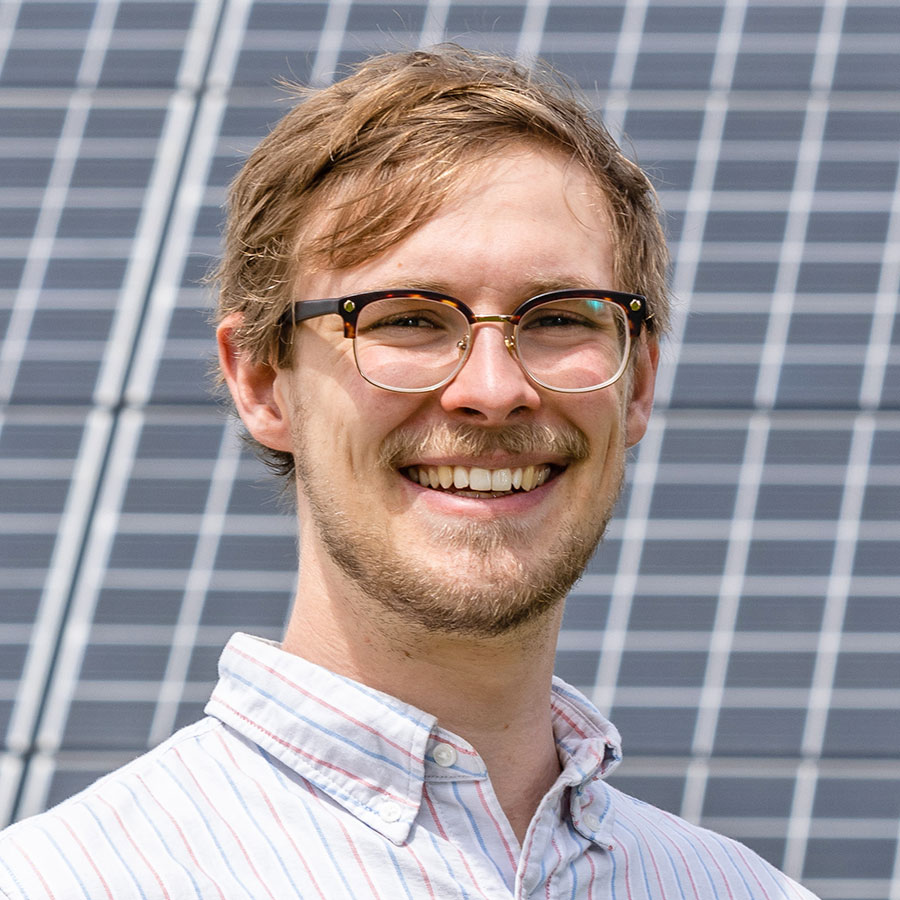 Blond man with beard and glasses, smiling, in front of solar panels