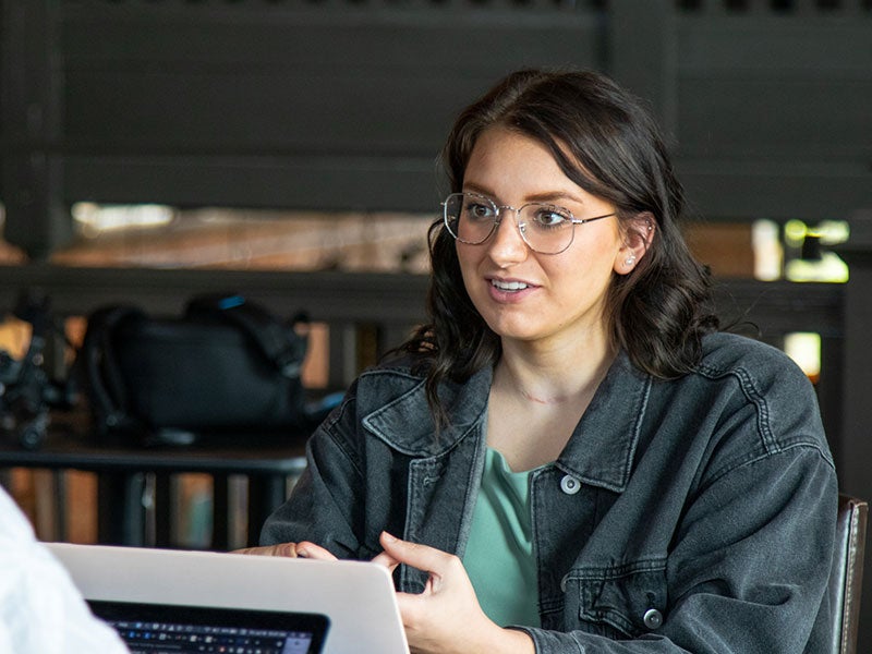 woman sits in a cafe with a laptop, talking to someone