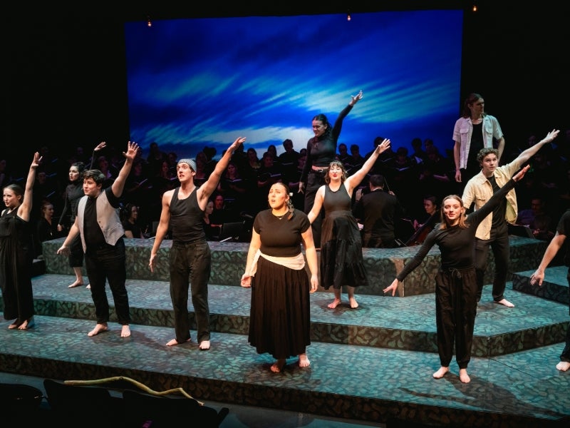 Actors in black clothing stand on a dramatically lit stage, singing.