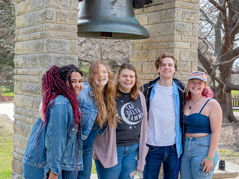 five happy Luther College students standing in front of the bell on campus