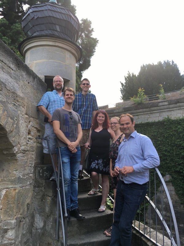 Six people stand on a curved stairway next to a stone wall. 