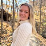 Sadie stands on a forest path, wearing a white sweater. 