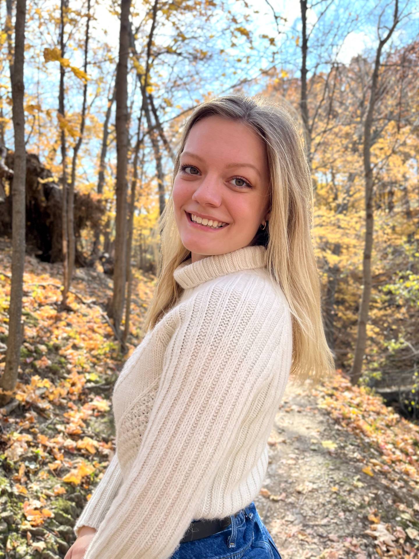 Sadie stands on a forest path, wearing a white sweater.