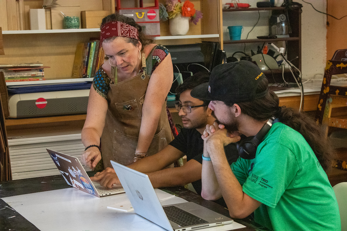 In the art center, two students sit at a desk with computers, looking at the ArtHaus director.