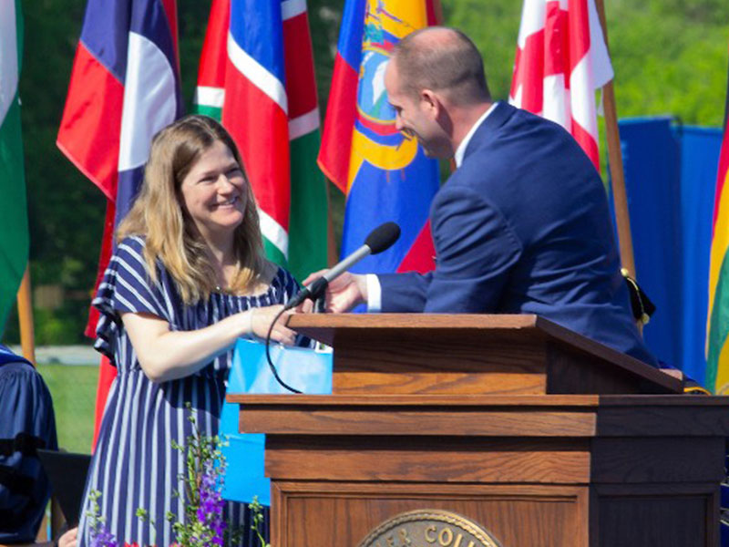 A woman on a stage in front of a row of flags shaking hands and accepting a gift bag from a man behind a lecturn