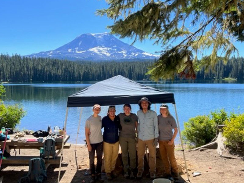 A group of five people standing under a tent at the side of a mountain lake