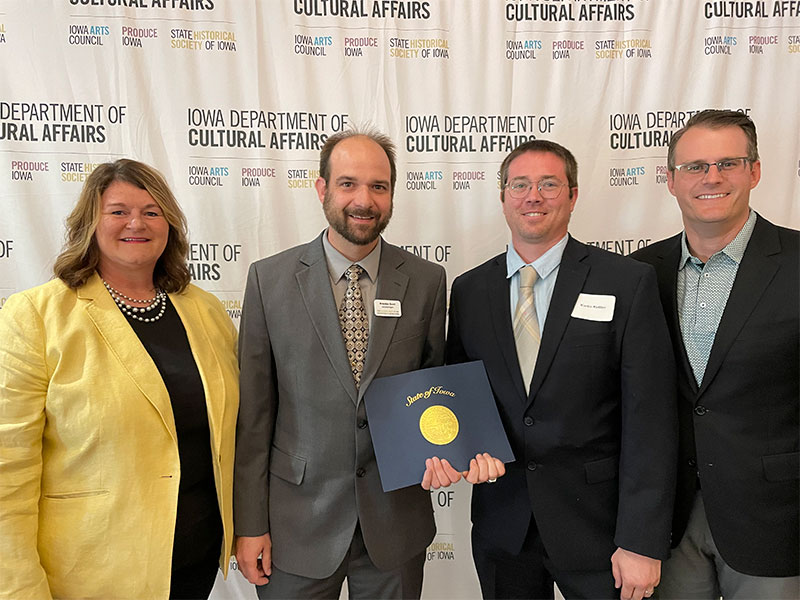 Four people, two holding an award, pose in front of a press backdrop