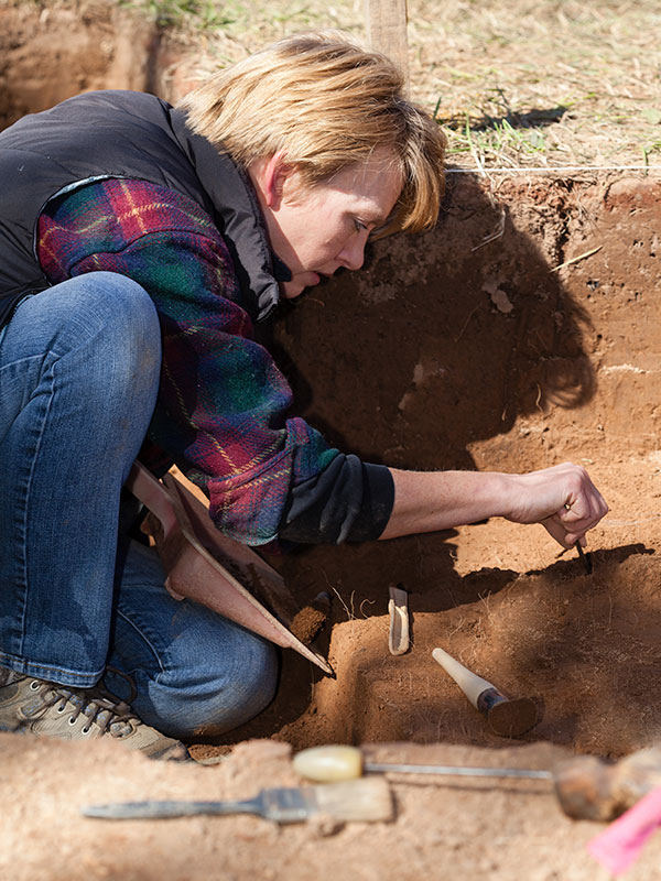 A woman crouching in an archaeological pit, working with intricate tools