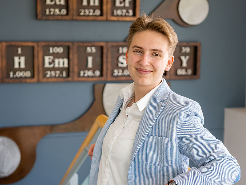 A woman leaning against a railing in front of a wall with the periodic table of elements on it
