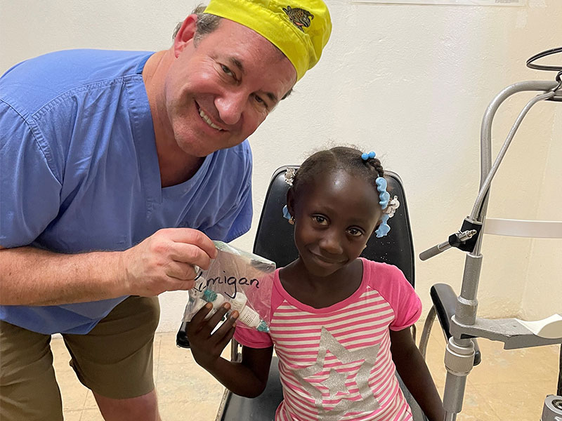 A man in a surgeon's cap bending over to give a girl in a medical chair some eye drops