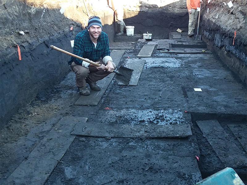 A man crouching in a straight-edged rectangular archaeological pit
