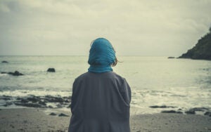 A woman wearing a gray coat and a blue head scarf stares at an unidentified sea.