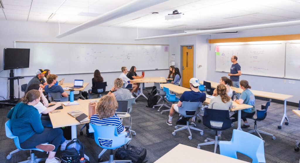 Students in a classroom sit around tables, having a discussion. 