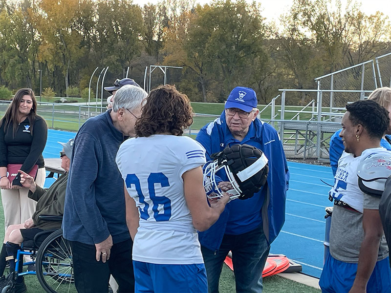 Two younger men in football uniforms talk with two older men on a football field