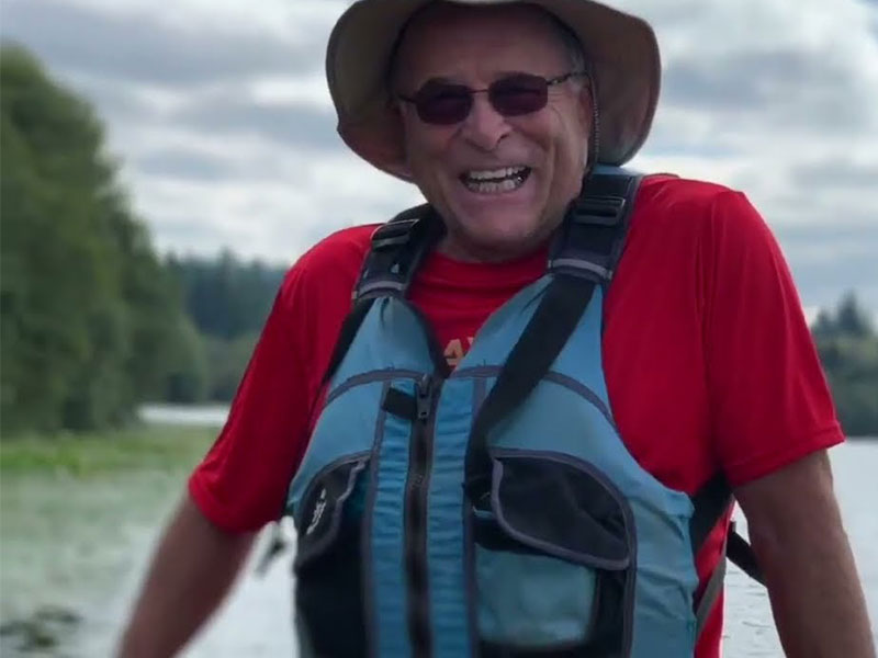 A man in a red T-shirt and blue life vest smiles in front of a backdrop of water and bluffs