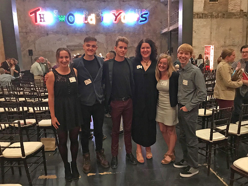 A group of six people pose indoors with a neon sign in the background