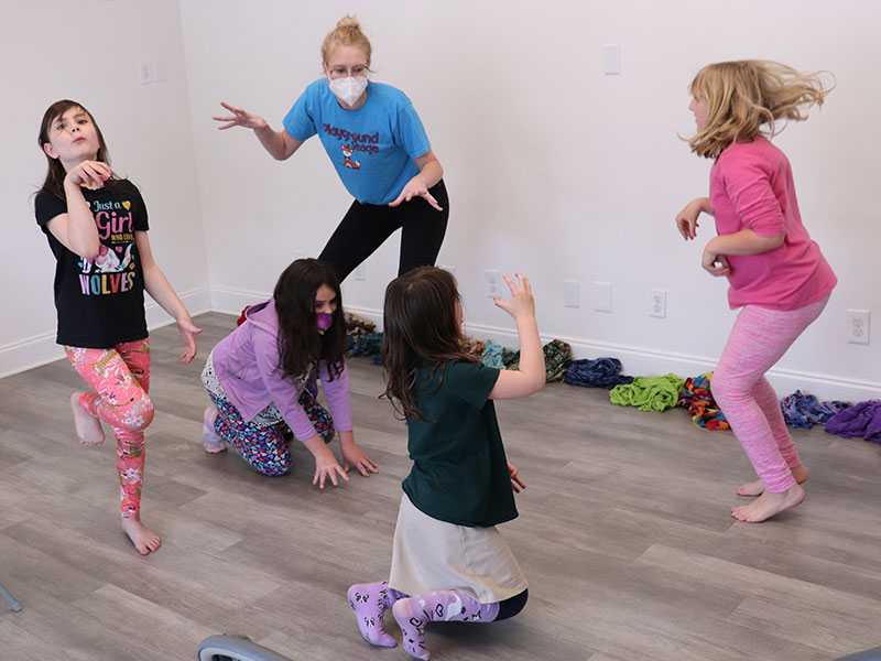 An adult woman wearing a mask poses crouches with arms posed like claws among a group of four children in similar dramatic poses