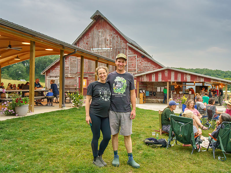 A woman and a man standing arm-in-arm in front of a covered patio and red barn, with people in lawn chairs in the background