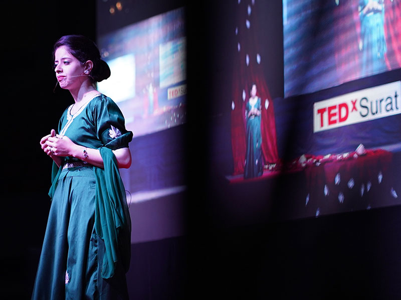 A woman wearing a headset on a dark stage with a TedXSurat backdrop