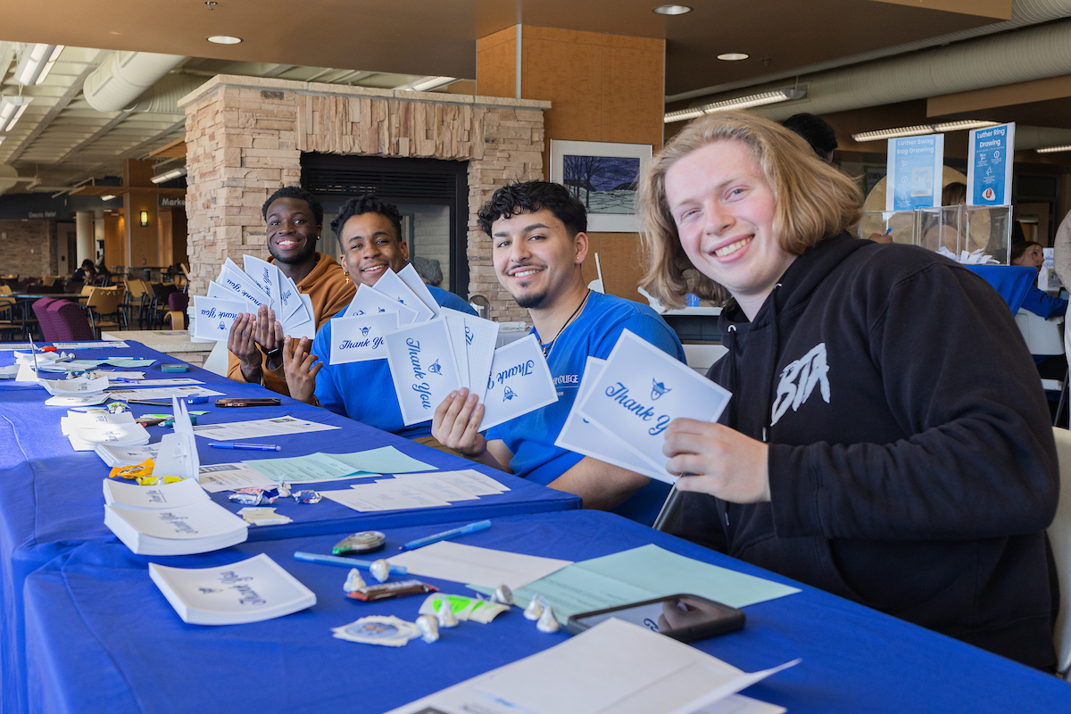 Students sit at a table with a blue tablecloth, smiling and showing off their thank you notes.