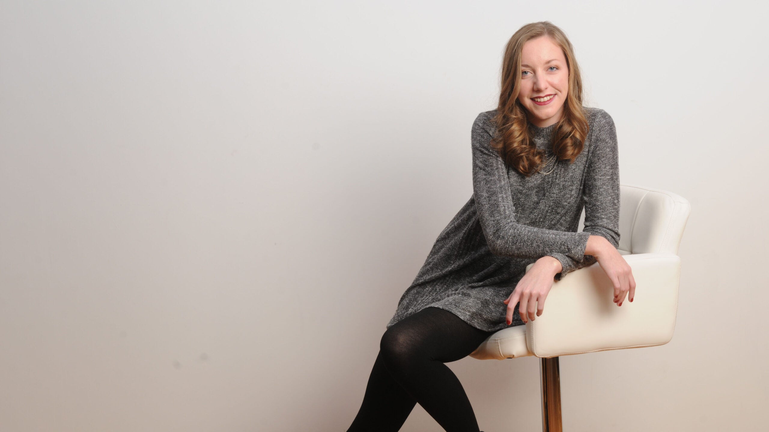 Wearing a gray dress, Kirsten sits on a white chair.