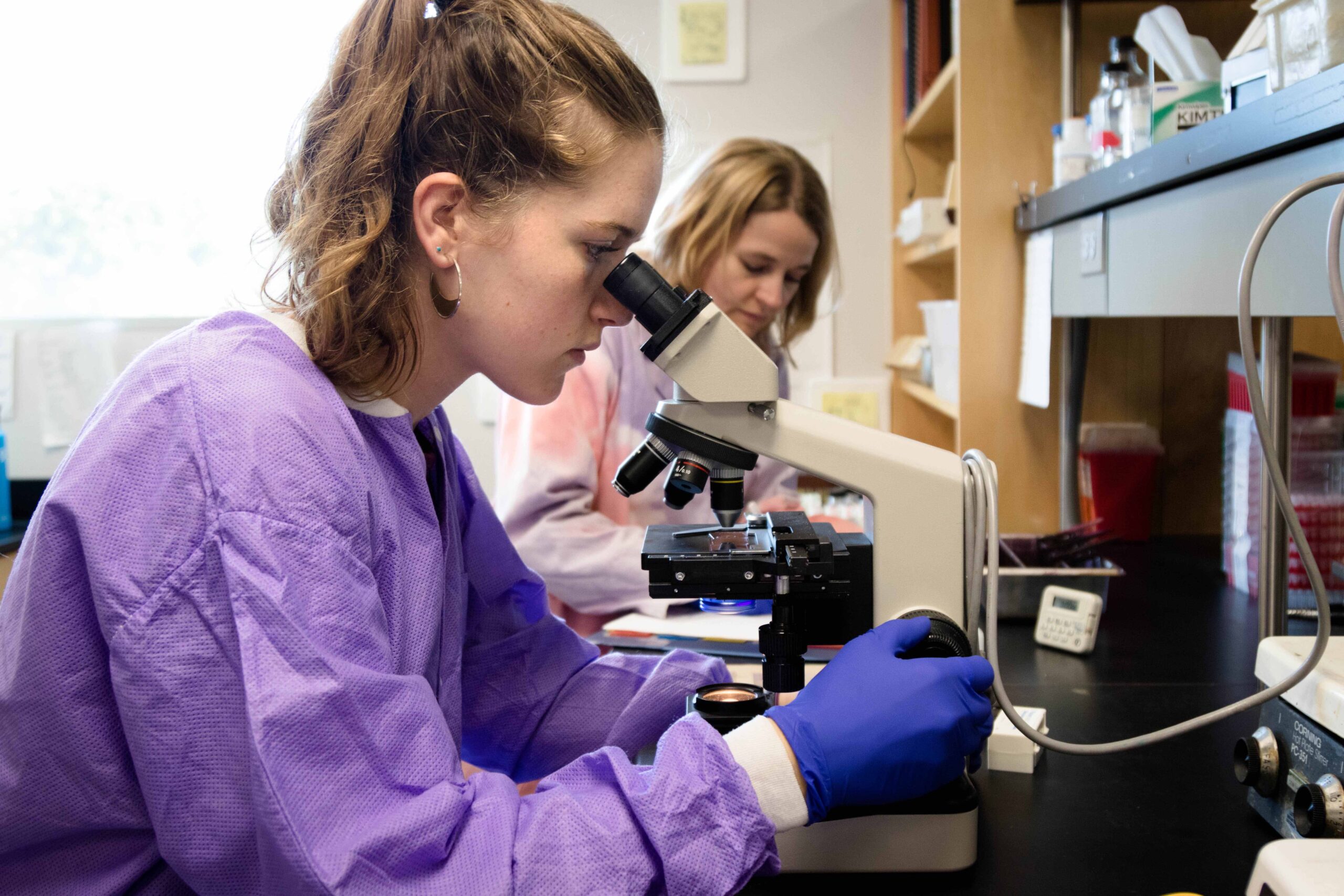 A student in purple lab coat and purple gloves peers into a microscope