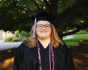 Matilda Koeller stands in front of a tree in a black graduation gown and cap.
