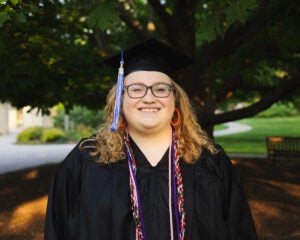 Matilda Koeller stands in front of a tree in a black graduation gown and cap. 