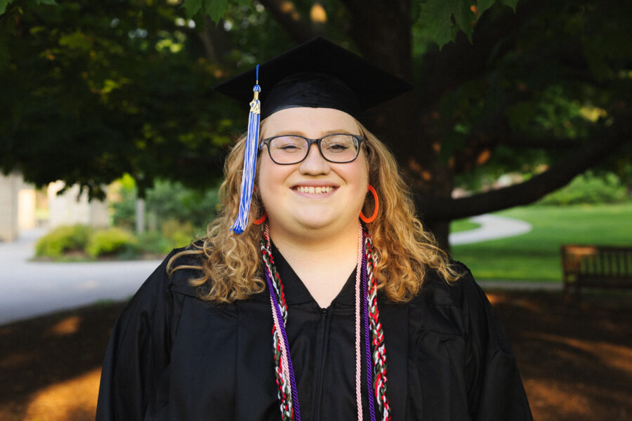 Matilda Koeller stands in front of a tree in a black graduation gown and cap.