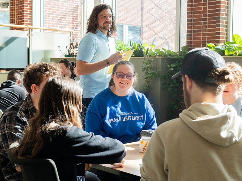 A man stands near a table of seated college students.