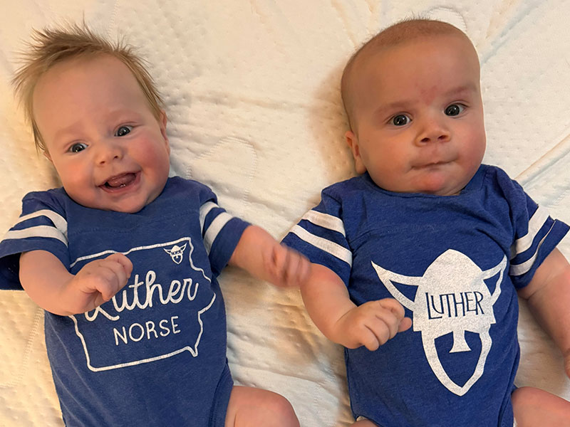 Two babies lying on the ground, wearing blue Luther College onesies