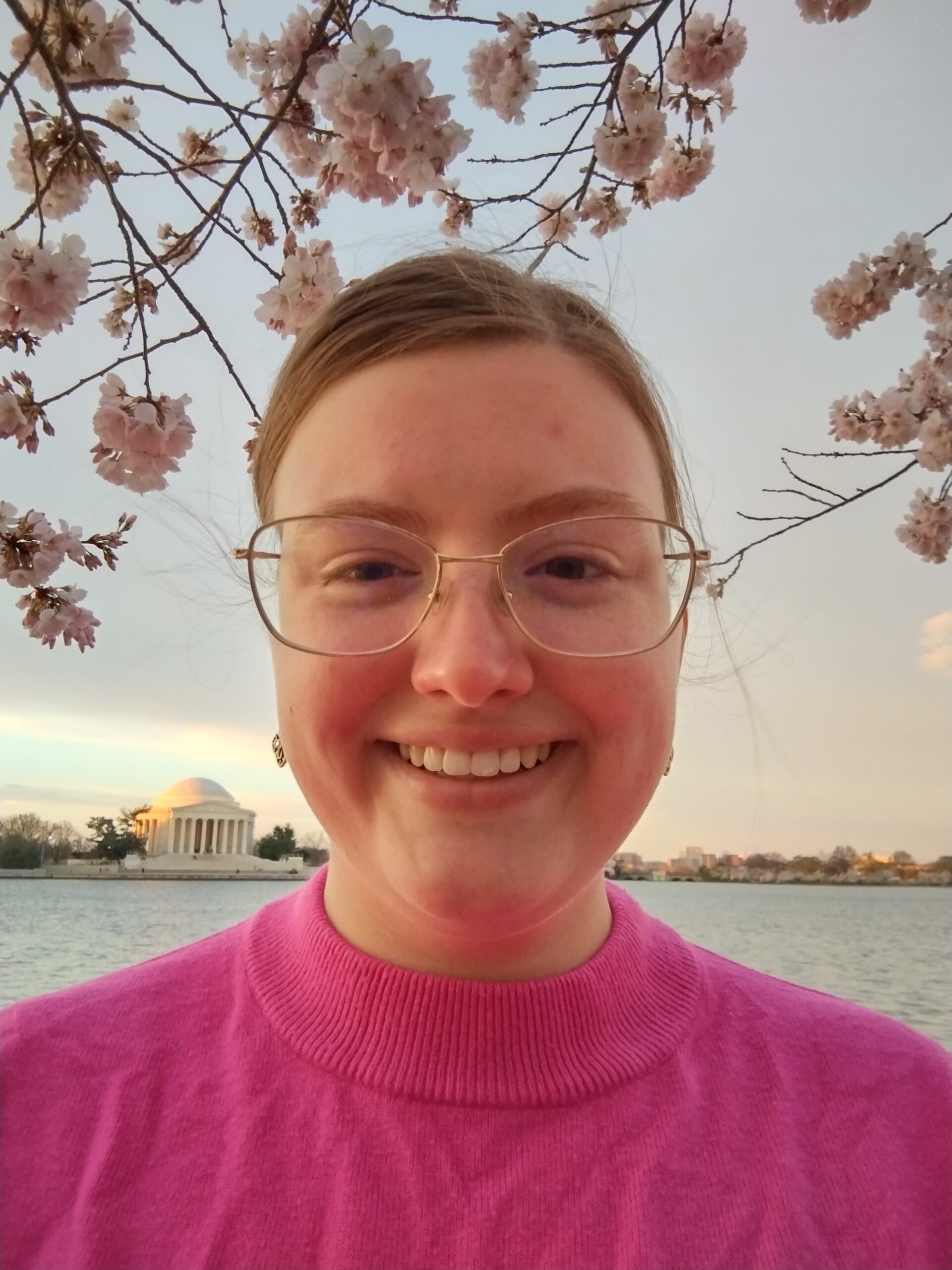 Emily Hursh in glasses and a pink sweater against a sunset on the water.