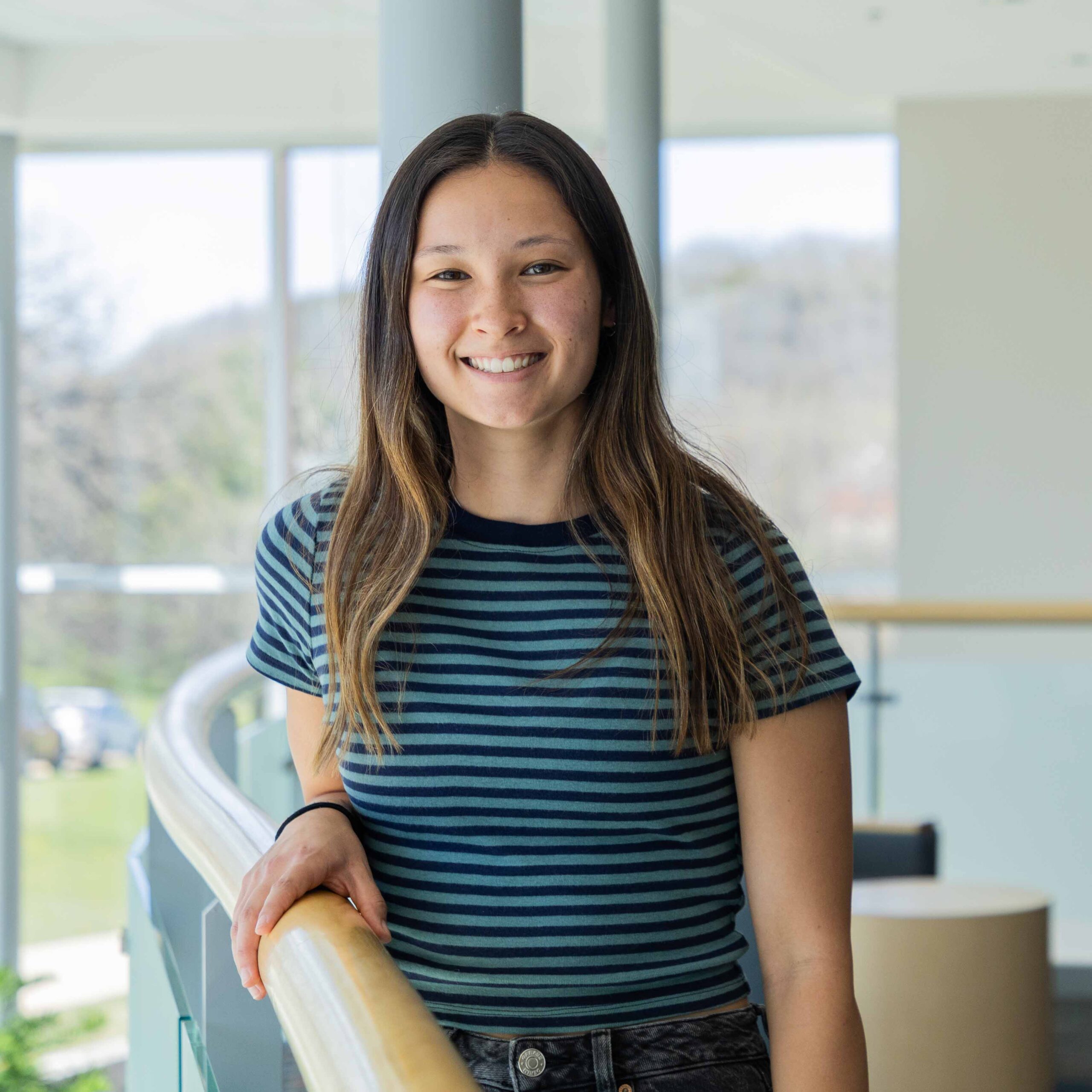 Kiley wears a blue striped shirt, stands in an atrium holding a wooden railing.