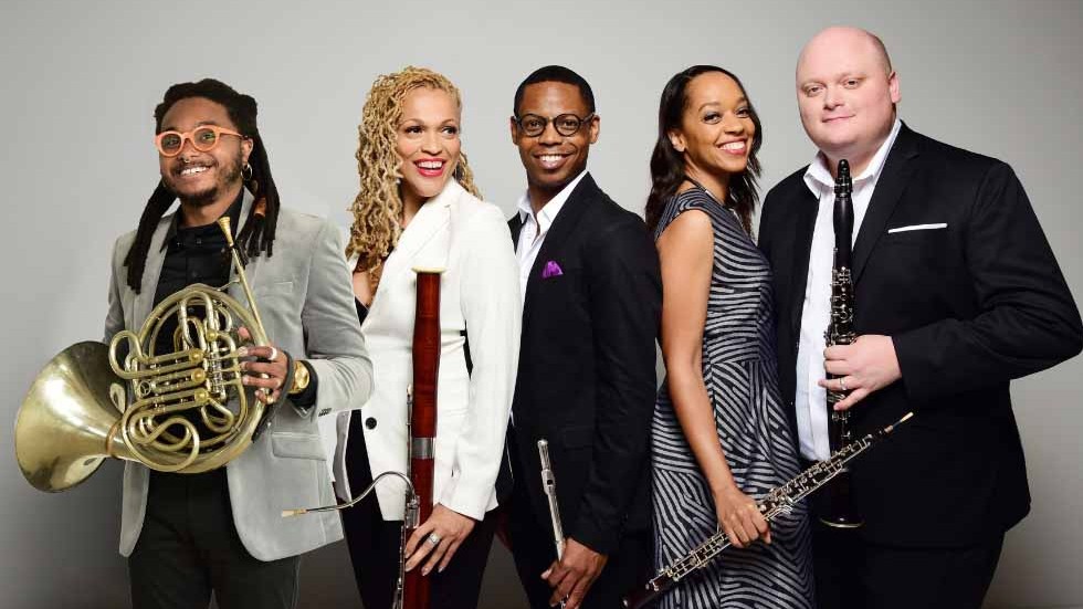 Members of Imani Winds holding their instruments in front of a grey backdrop