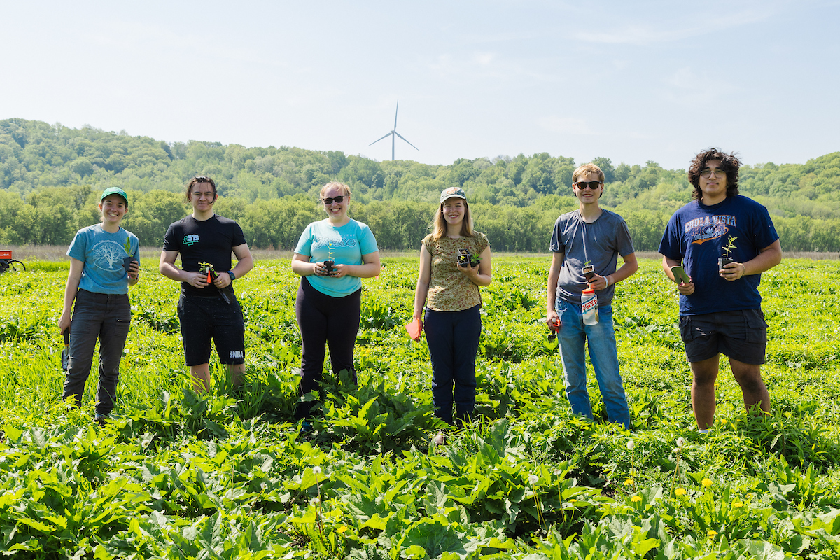 Six young people stand in a prairie with a wind turbine in the distance.