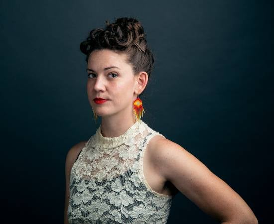 Rebecca Nagle, portrait with grey background, hair in an up-do, and dangly orange-yellow earrings.