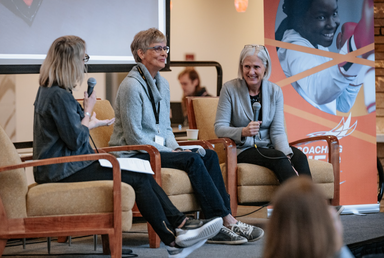 Three women sit in chairs on a raised platform, addressing an audience