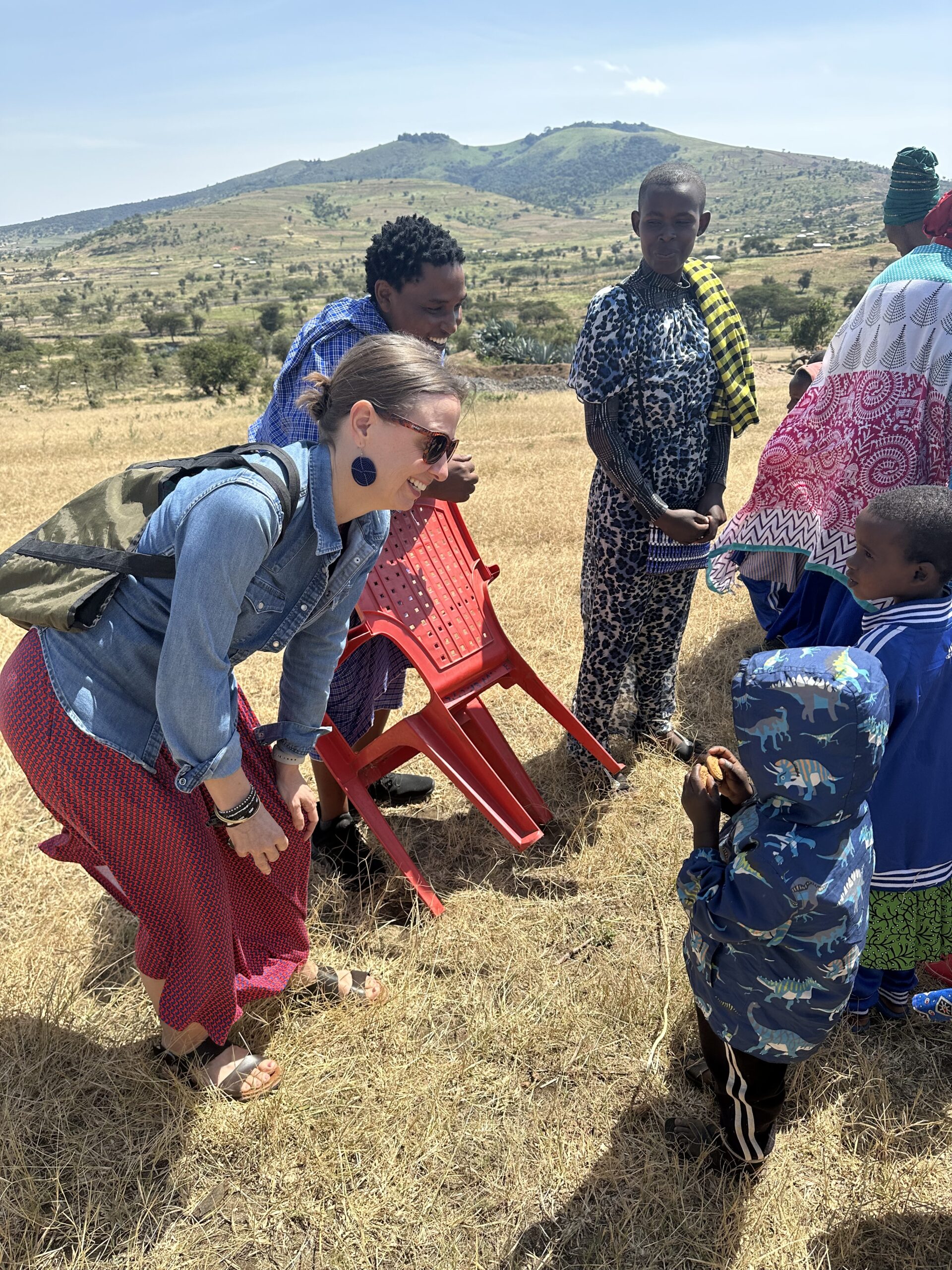 A casual circle of people, one holding a red plastic chair, gather in a sunny scrubland