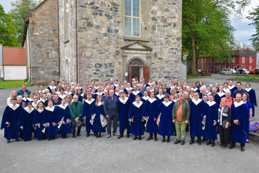A large group of people, most in blue choir robes, stand in front of an old, large stone building